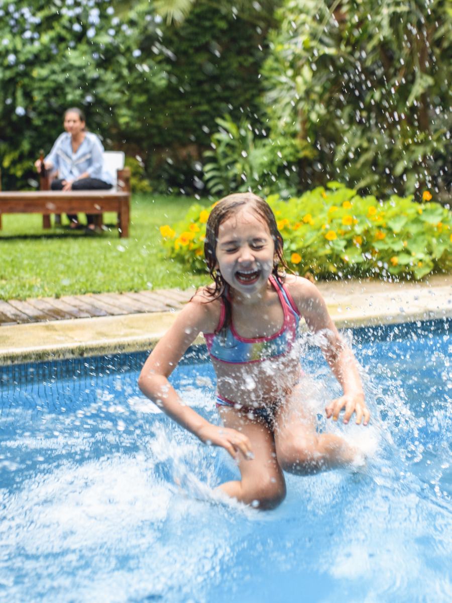 A child leaping into a clean backyard pool while two adults watch from a shaded bench